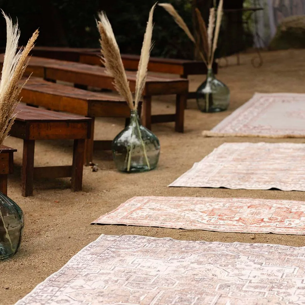 Wooden ceremony benches at Torre Sever, with glass demijohns holding pampas grass and patterned rugs on the sand