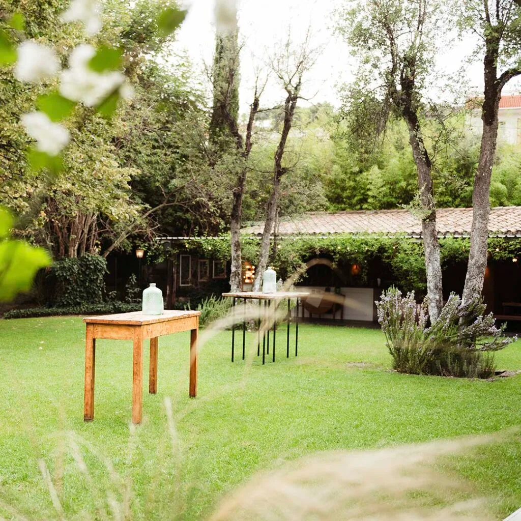 Garden aperitif area at Torre Sever with small wooden tables and bottles on the lawn, mature trees, and the stone arcade behind