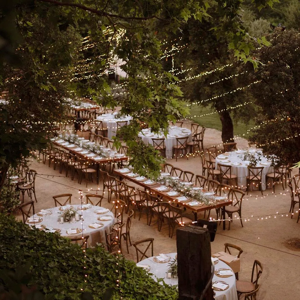 Outdoor dinner setting at Torre Sever with laid round and long tables, wooden chairs, and strings of lights among the trees