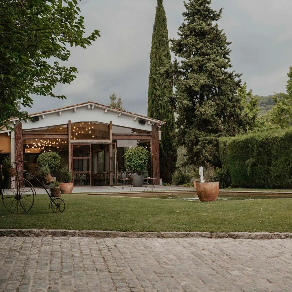 Exterior of the Torre Sever reception salon with open glass doors, warm string lights inside, lawn, fountain, cypress, and hills beyond