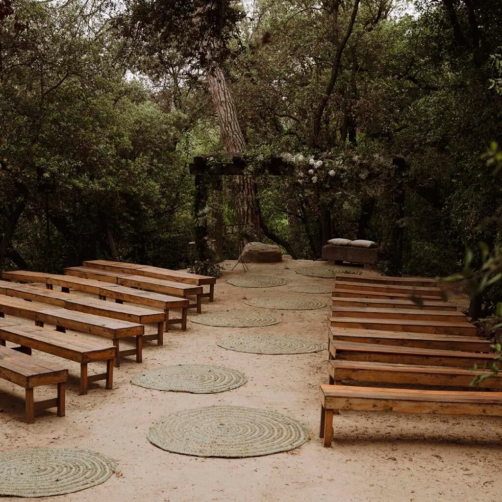 Woodland ceremony clearing at Torre Sever with wooden benches along a sandy aisle, woven rugs, and a flower-decorated timber structure